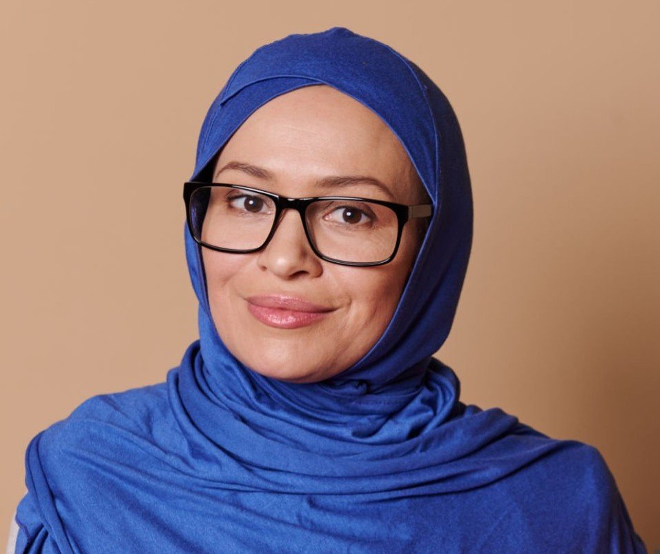 Confident portrait of a beautiful middle-aged Middle-Eastern Muslim woman, a teacher educator at high school, in blue hijab, carrying stack of books and looking at camera, isolated on beige background